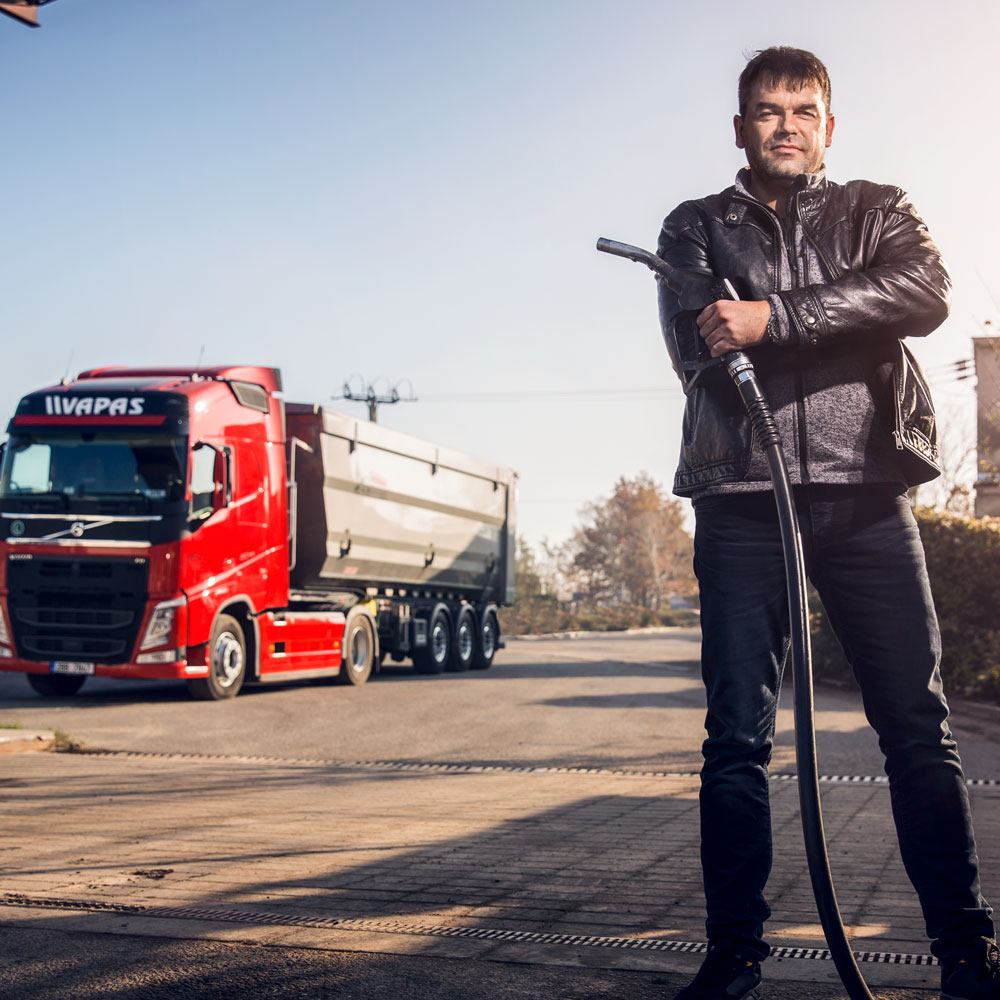 Petr Šišlák se encuentra en primer plano, sosteniendo un surtidor de gasolina con un camión Volvo detrás de él.