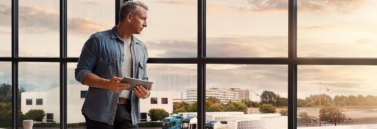 Un hombre que sostiene una tableta está junto a una ventana y mira hacia abajo su flota de camiones
